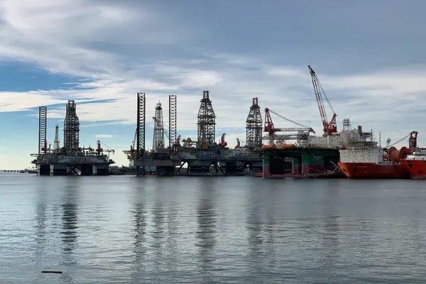 Jack-up drilling rigs and a red and white supply vessel line the far side of Galveston harbor under a cloudy sky, the channel water flat in the foreground