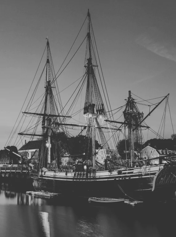A three-masted tall ship tied up at a Salem wharf at dusk, its rigging webbed against the sky and a rowboat floating alongside