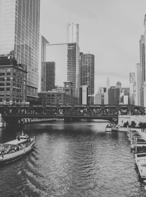 A tour boat pushes up the Chicago River in black and white, passing beneath an elevated rail bridge with downtown towers stacked behind it