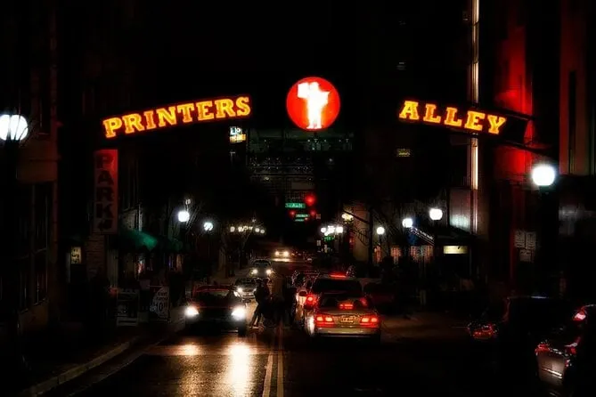 A red and gold neon Printers Alley sign arches over a dark downtown Nashville street at night, headlights and taillights crowding the wet pavement below