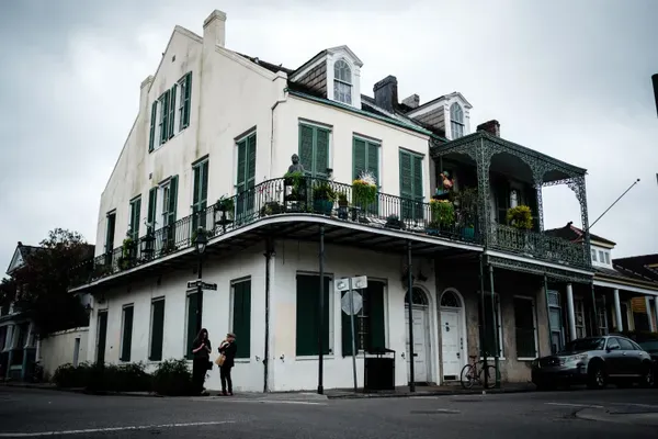 A white corner building in the French Quarter with green shutters and two levels of iron-lace balconies crowded with potted plants, two people talking on the sidewalk below the street sign