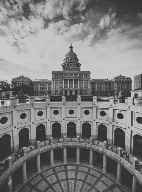 The Texas State Capitol dome rises beyond the open-air rotunda of its underground extension, a ring of arches and columns in the foreground under streaked clouds, in black and white