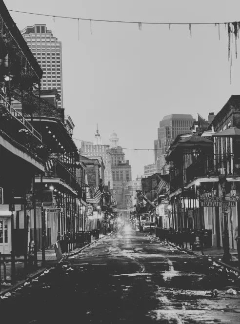 A French Quarter street in black and white lined with cast-iron balconies and bar signs, strands of beads hanging from the overhead wires, downtown towers rising in the haze at the far end