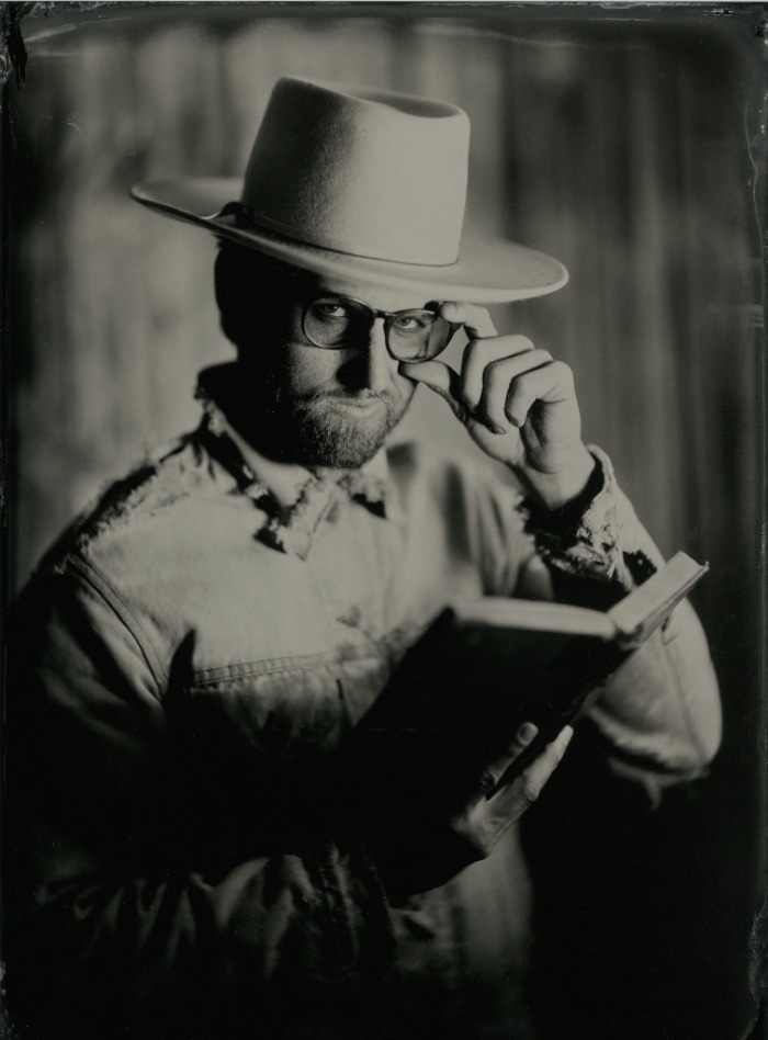 Tintype-style portrait of a bearded man in a wide-brim hat holding an open book