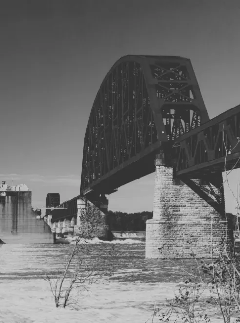 A steel railroad bridge on stone piers crosses the Ohio River in black and white, whitewater spilling over a low dam beyond it