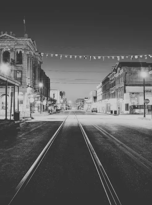 Rail tracks run down the middle of an empty Galveston street at night between a columned stone building and a row of lamplit storefronts, pennant flags strung overhead, in black and white