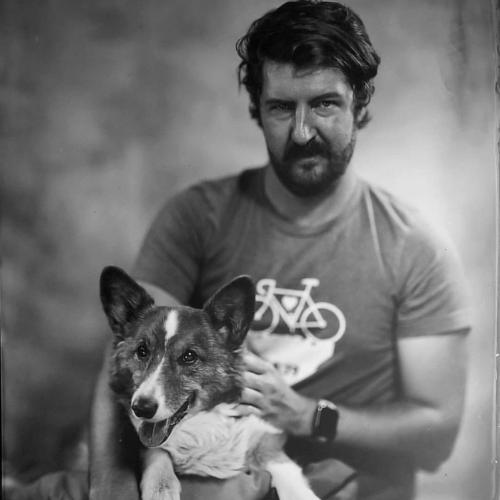 Black-and-white portrait of a bearded man in a bicycle T-shirt holding a dog