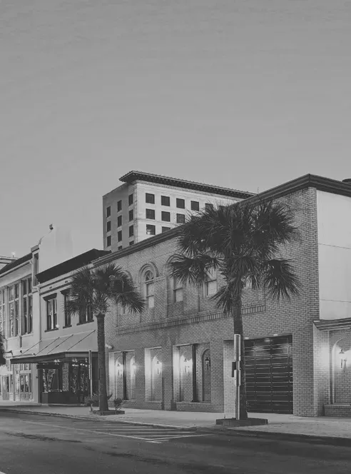 A two-story brick commercial block with arched upper windows and lit shopfronts stands behind palm trees on an empty Galveston street at dusk, in black and white
