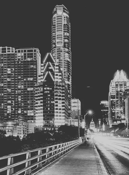 Night on a bridge walkway into downtown Austin, glass towers lit above the guardrail, the Capitol dome small and bright between them, car lights streaking down the roadway, in black and white