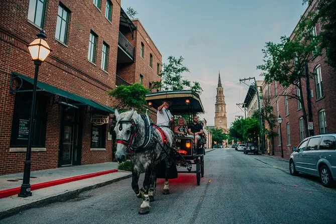A dappled gray draft horse pulls an open carriage of seated riders down a brick-lined Charleston street at dusk, a white church spire closing the view ahead