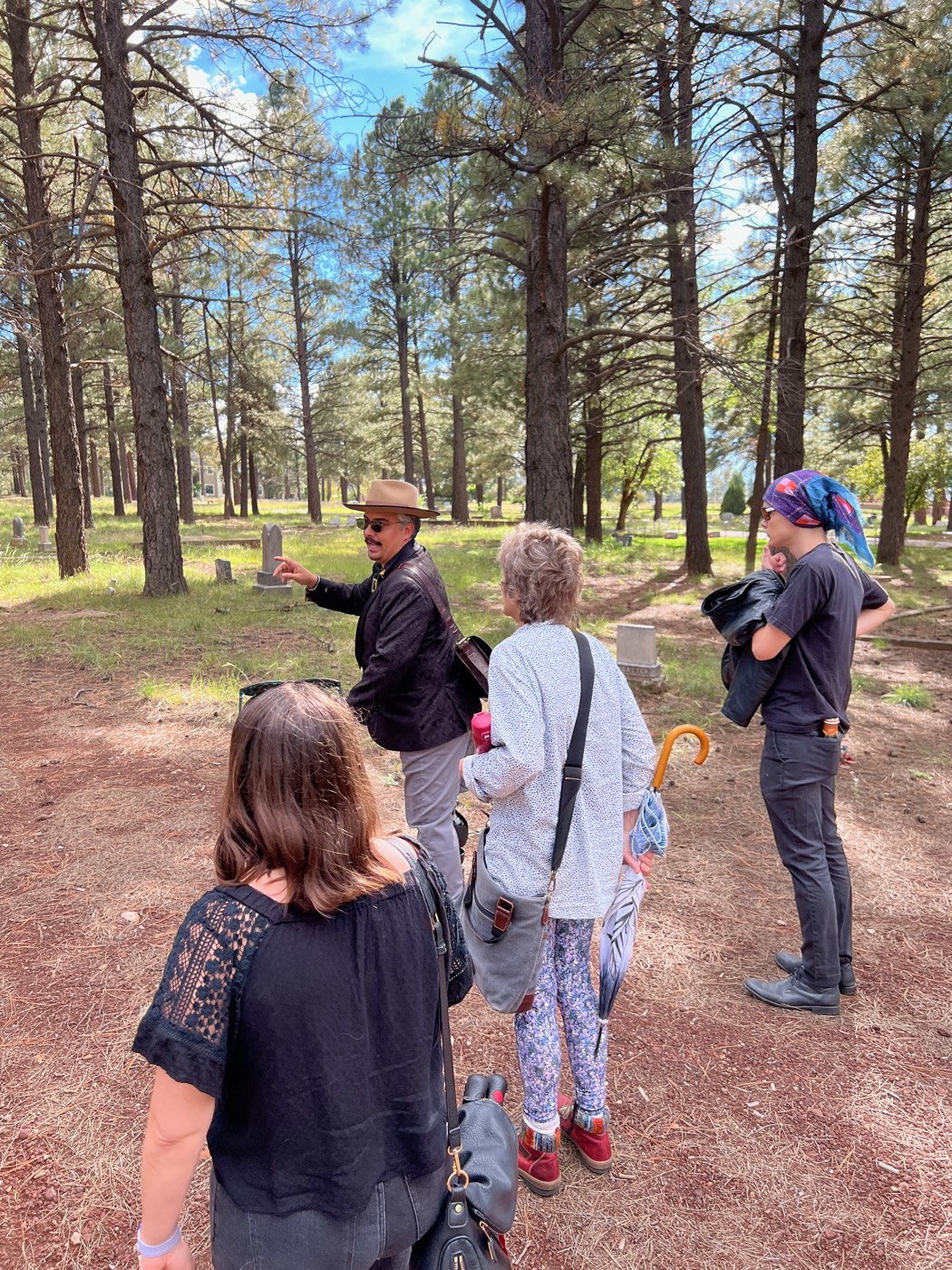 Deep among the pines of Citizens Cemetery, a guide points out a weathered headstone to guests carrying umbrellas against the monsoon sun on Flagstaff's cemetery walking tour.