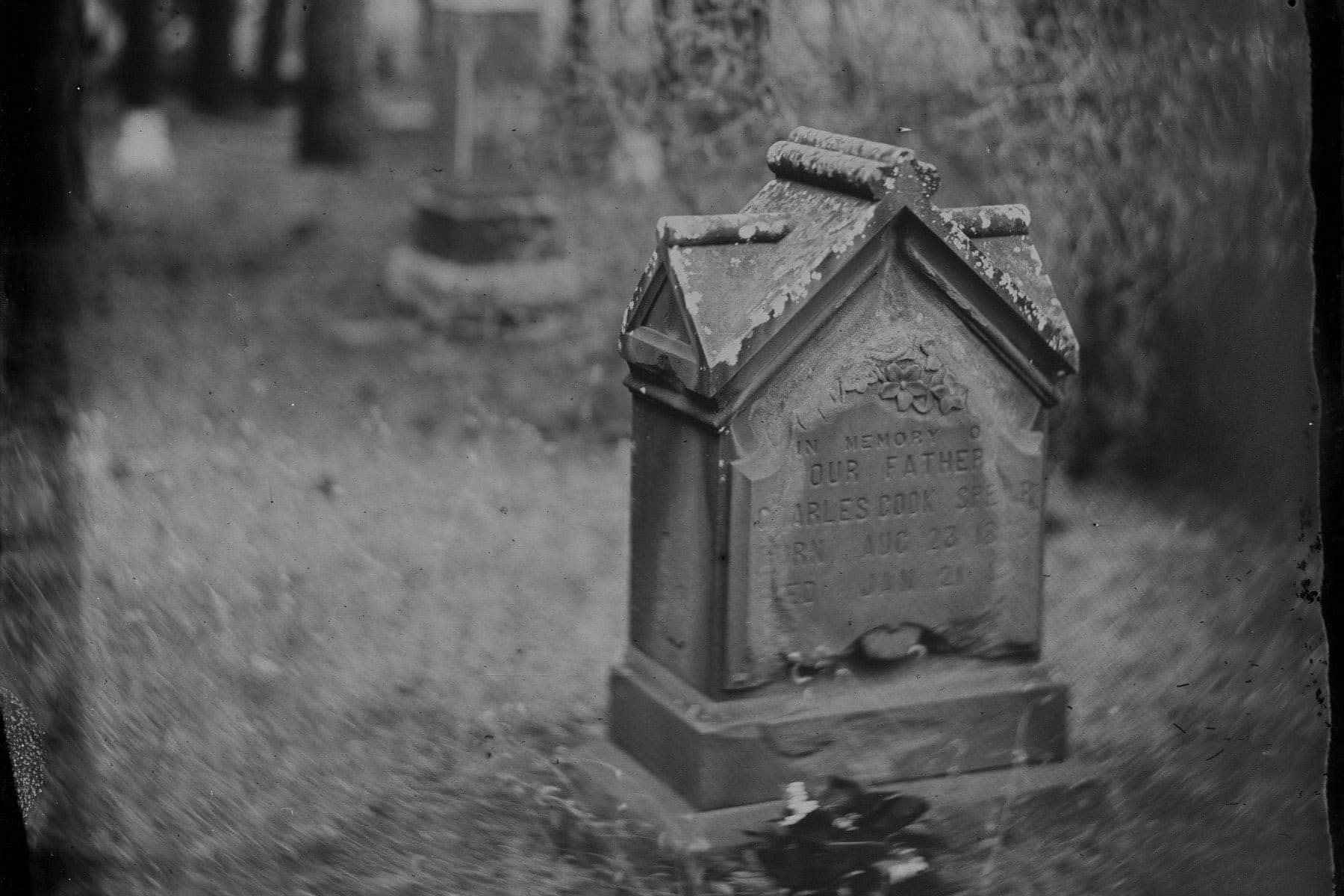 A weathered peaked grave marker stands among the pines at Citizens Cemetery, older headstones soft in the distance.
