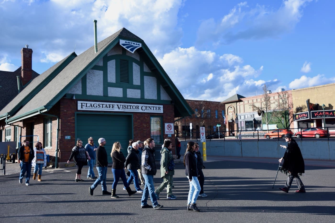 Guests file past the Tudor Revival gable of the Flagstaff Visitor Center, still an Amtrak depot, while their caped guide waits with a cane on the old Route 66 alignment.
