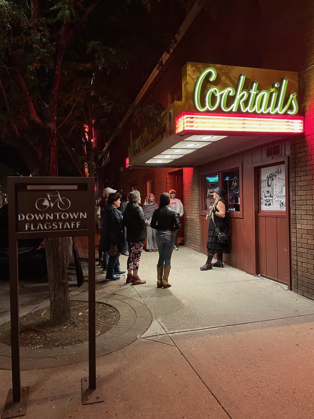 The green Cocktails neon of the Monte Vista Lounge burns over a nighttime tour group beside the Downtown Flagstaff sign, a stop on the 21-and-over Spirits With The Spirits haunted pub crawl.