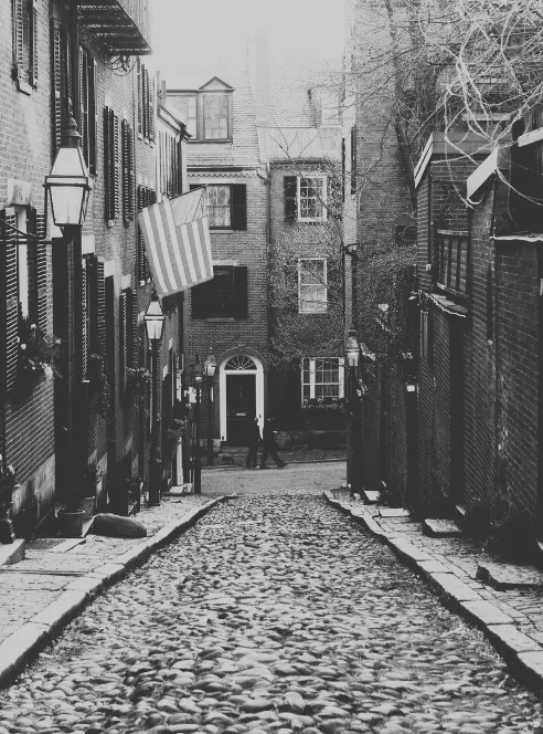 A steep cobblestone lane between brick row houses with black shutters and gas lamps, an American flag hanging over the sidewalk and a lone figure crossing at the far end, in black and white