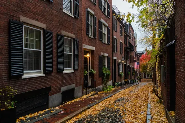 Yellow autumn leaves cover the cobblestones of a narrow lane lined with brick row houses, black shutters, window boxes, a lit doorway and a small American flag further down