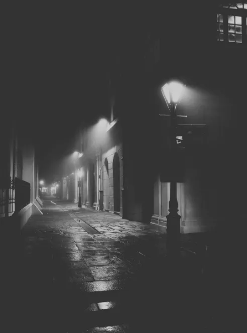 A narrow French Quarter street at night in black and white, a single gas lamp burning on its post above wet flagstone paving, shuttered arched doorways receding into the dark