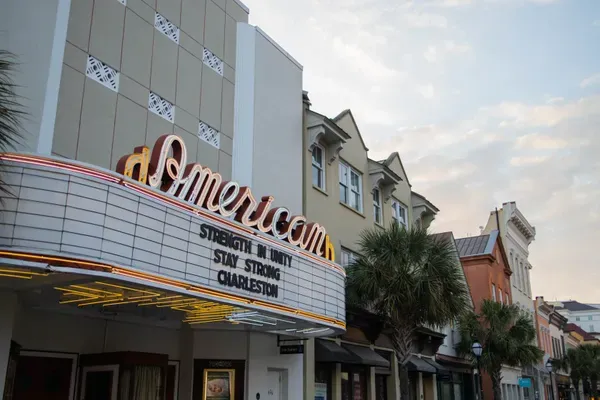 The American Theater's neon marquee glows over a King Street sidewalk at dusk, its letter board spelling out STRENGTH IN UNITY STAY STRONG CHARLESTON beside a palmetto tree