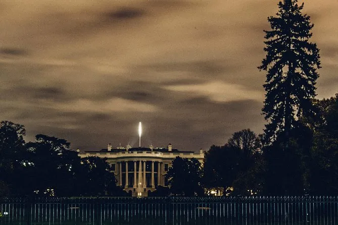 The White House lit behind a long iron fence at night, dark trees on either side and a flagpole catching the light above the roofline
