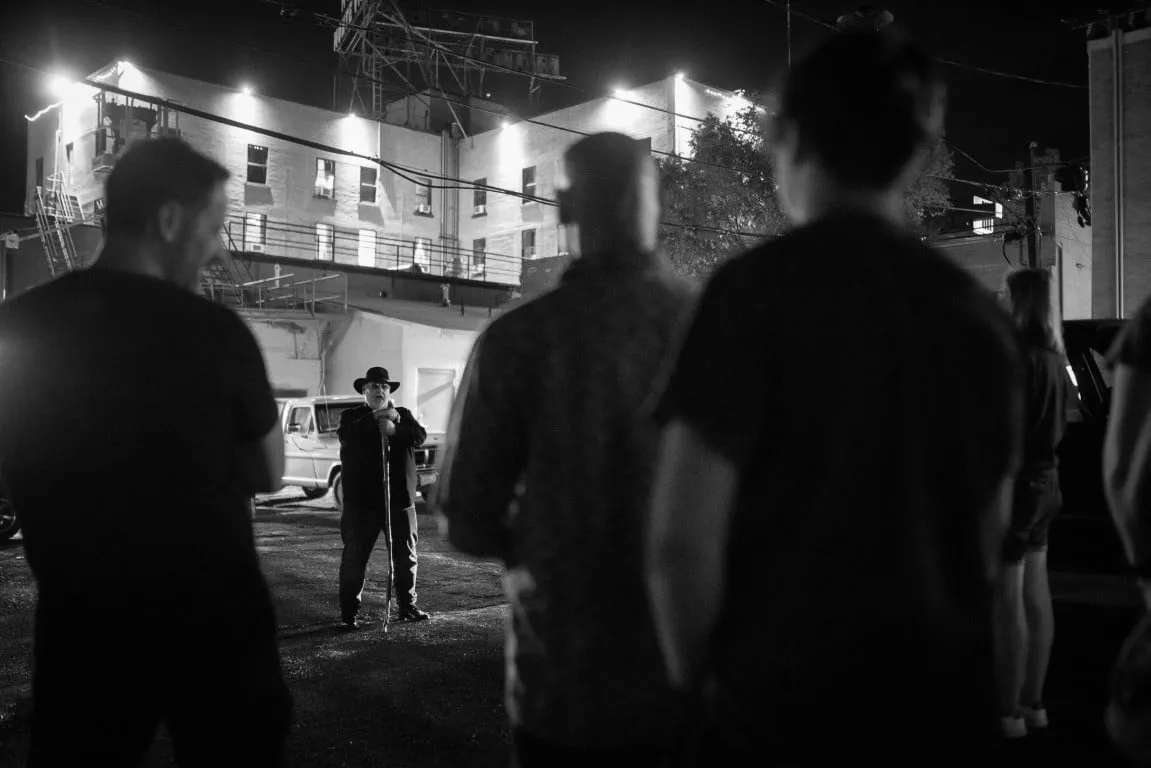 Black and white night scene of a guide addressing a tour group outside a lit building