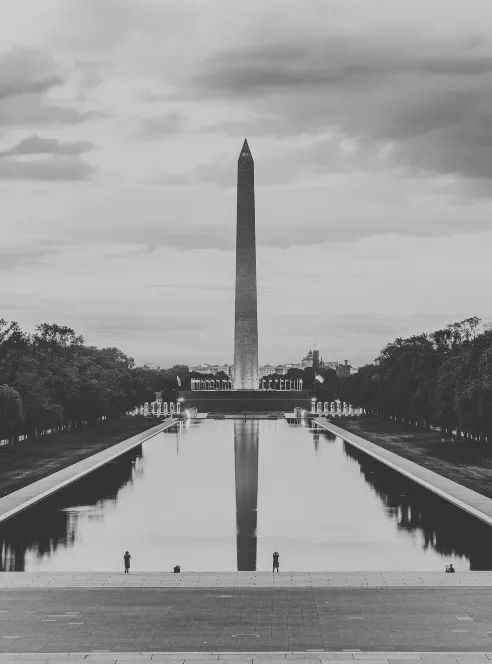 The Washington Monument mirrored full length in the Reflecting Pool at dusk, two small figures on the far pavement, in black and white