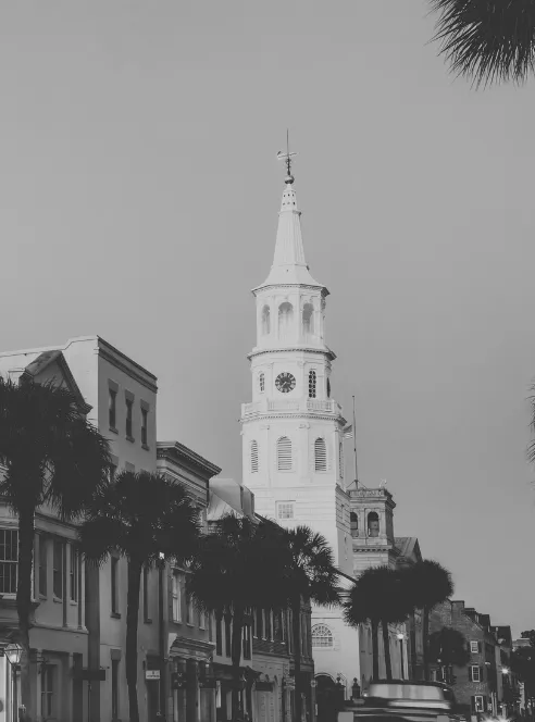 A white church steeple with a clock face rises above a row of Charleston storefronts and street palmettos, photographed in black and white