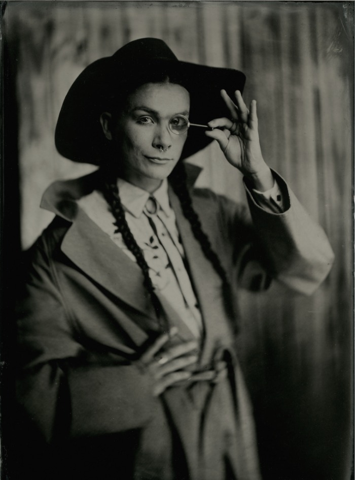 Tintype portrait of a person in a hat and suit adjusting a monocle, with long braids
