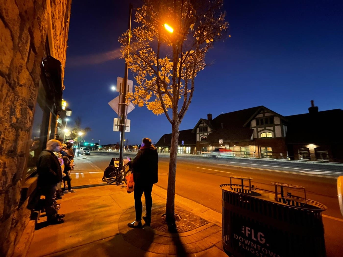 A tour group on a dark Flagstaff sidewalk with the train depot lights in view